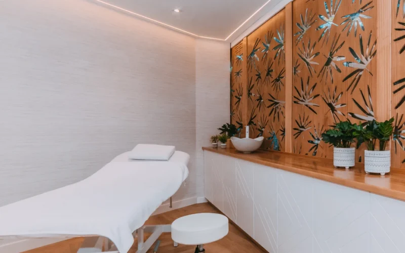 Proyecto Wellness Parque Santiago 3. Una sala de spa minimalista con camilla de masajes, paneles decorativos de madera, plantas e iluminación tenue.